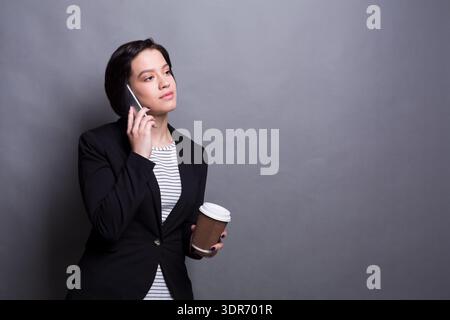 Businesswoman Talking on the Phone While Holding a Coffee Cup in Front of a Gray Background Stock Photo
