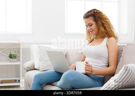 Woman Working on a Laptop While Sitting on a Couch With a Cup in a Bright Room Stock Photo