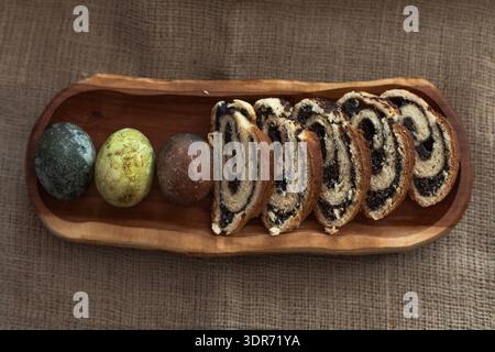Sliced homemade poppy seed roll and three colorful Easter eggs arranged on a wooden plate, rustic holiday composition Stock Photo