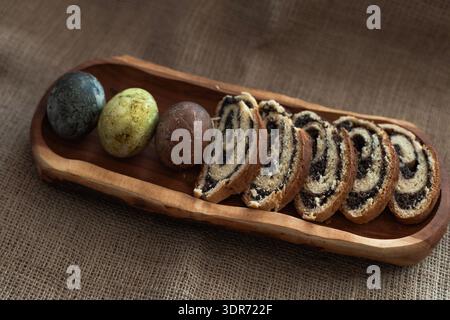 Traditional Easter composition with sliced poppy seed roulade and three dyed eggs on wooden serving board Stock Photo