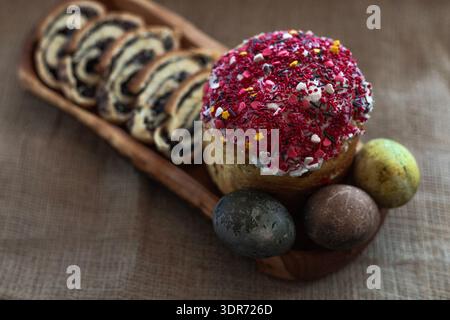 Easter still life with sliced poppy seed roll, three dyed eggs and traditional Easter cake on a wooden plate, rustic festive setting Stock Photo