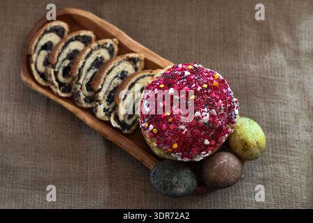 Flat lay composition of homemade Easter bread topped with icing, three dyed eggs and sliced poppy seed roulade on a long wooden tray, traditional Stock Photo