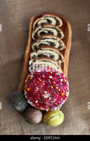 Overhead shot of three colorful Easter eggs, decorated Easter cake with icing and sliced sweet roll served on elongated wooden plate, cozy rustic back Stock Photo