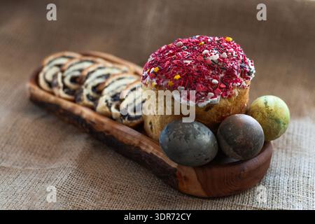Top view of traditional Easter table with three dyed eggs, homemade glazed Easter bread and sliced poppy seed roll arranged on a long wooden serving b Stock Photo