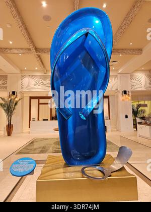 EDITORIAL USE ONLY- MARGARITAVILLE RESORT HOLLYWOOD BEACH FLORIDA: ​A giant, glossy blue flip-flop sculpture titled "Blown Out Flip Flop" stands as a centerpiece in the lobby of the Margaritaville Hollywood Beach Resort in Hollywood, Florida. The pop-art sculpture features a broken strap and is accompanied by a large metallic pop-top "tab" resting on a golden, wave-textured base. The background showcases the resort's bright, coastal-themed interior with white-washed wooden beams, tropical palm plants, and elegant white architectural details. This iconic art installation is a playful reference  Stock Photo