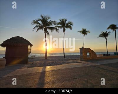 EDITORIAL USE ONLY- HOLLYWOOD BEACH FLORIDA: : A vibrant sunrise at Hollywood Beach, Florida, featuring a traditional thatched-roof tiki hut in the foreground. The sun peeks from behind a silhouetted palm tree, casting long shadows across the brick-paved Broadwalk. The scene includes decorative masonry architecture and a row of palm trees lining the Atlantic shore, perfectly capturing the tropical, "Old Florida" charm of this popular vacation spot at the start of a clear day. Stock Photo