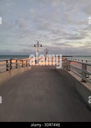 EDITORIAL USE ONLY- KEY WEST FLORIDA:  A wide-angle, symmetrical perspective of the Edward B. Knight Pier (White Street Pier) during twilight in Key West, Florida. The concrete promenade is illuminated by warm lamp posts, casting orange light on the ground against a backdrop of soft blue evening clouds and the Atlantic Ocean. Two distant figures are seen walking along the vast, peaceful structure. Stock Photo