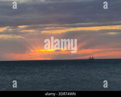 EDITORIAL USE ONLY- KEY WEST FLORIDA:  Scenic coastal landscape at dusk in Key West, Florida, featuring a traditional sailing schooner silhouetted against a vibrant orange horizon. Dramatic crepuscular rays (sunbeams) radiate upward through a break in the heavy grey cloud cover. The calm waters of the Gulf of Mexico fill the foreground under a moody, atmospheric tropical sky. Stock Photo