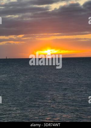 EDITORIAL USE ONLY- KEY WEST FLORIDA:  A vibrant tropical sunset over the Gulf, captured from Key West, Florida. The bright sun disk sinks toward a flat horizon line, framed by dramatic dark grey and orange-rimmed clouds. The deep blue water of the Florida Keys shows gentle surface ripples reflecting the intense golden hour glow. A distant maritime marker is visible on the horizon, emphasizing the vast coastal seascape. Stock Photo