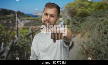 Young man with beard wearing white t shirt and fist extended toward camera in forest path; determination grit resilience. Stock Photo