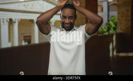 Man raises hands above head showing forearms and smile, wearing white polo, facing camera on old town building balcony with classical columns; confide Stock Photo