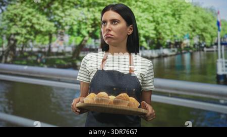 Woman baker holding tray of muffins with both hands on a street by a canal, presenting pastries; curious hospitality. Stock Photo