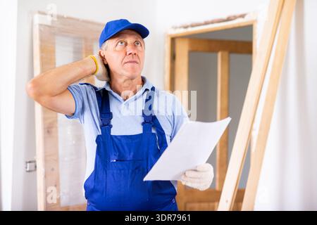Worried middle-aged male builder in a blue uniform holding home construction blueprint during repair indoors Stock Photo