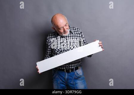 Older Man Holds Blank Sign While Standing Against Grey Background in Indoor Setting Stock Photo
