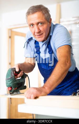 Focused middle-aged man in a blueuniform using a jigsaw machine on wooden plank at home Stock Photo