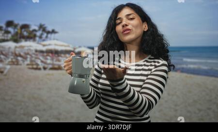 Woman holding moka pot and blowing a kiss toward camera in studio beach set while wearing striped top and smiling; playful greeting. Stock Photo