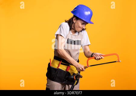 Female builder in hardhat and tool belt grips handsaw with focused expression while simulating cutting action. Renovation worker in studio demonstrates DIY effort against bold yellow background. Stock Photo