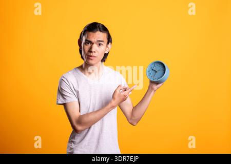 Concerned man holds alarm clock and gestures urgently toward time. Male individual looks uneasy and stressed, expressing awareness of deadlines, punctuality and rush of managing busy daily schedules. Stock Photo