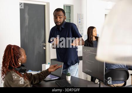 African american male and female coders collaborate at workplace, reviewing HTML scripts on desktop pc. Teamwork and focused communication support innovation in web and software development. Stock Photo