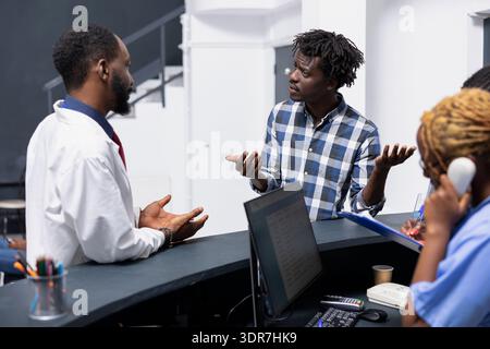 Young male adult gestures while discussing concerns with doctor at reception counter of medical clinic. The interaction highlights communication and coordination in patient support services in hospital Stock Photo