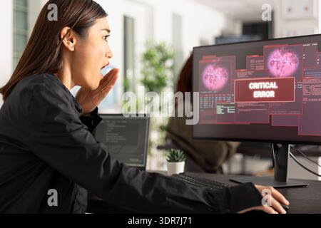 Asian female developer reacts in shock, covering mouth while staring at critical error message on computer screen, signaling system crash or cyberattack. Woman programmer panics over coding failure. Stock Photo