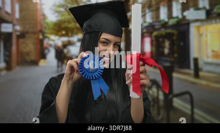 Young hispanic woman in graduation cap and gown proudly holds diploma and blue ribbon while standing on urban street. Stock Photo