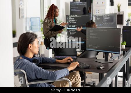 Asian programmer in wheelchair types code at office desk with focused concentration. Software developer analyzes backend data, representing accessibility, inclusion and innovation in technology. Stock Photo