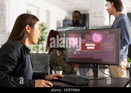 Asian female IT professional sits at desk staring at device screen displaying critical error message. Shocked reaction highlights hacking, system failure, internet security breach and workplace stress Stock Photo