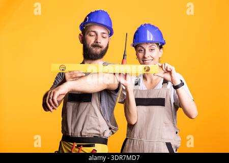 Male and female builders smile while holding measuring level together, demonstrating teamwork and accuracy at renovation site. Construction workers collaborating on building plans with tools in hand. Stock Photo