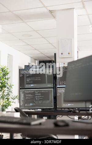 Vacant IT workspace features desk with multi monitors displaying coding scripts and system data. Empty office scene highlights innovation, software development and professional engineering workflow. Stock Photo