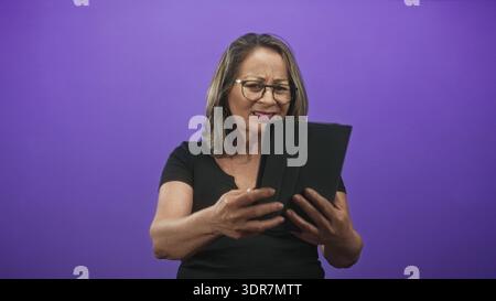 Hispanic woman with grey hair holding tablet with both hands, tapping screen and frowning in studio; digital confusion. Stock Photo