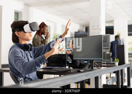 Male programmer uses VR glasses, interacting with 3D interface and analyzing coding scripts. Young male adult working at innovative company, exploring virtual technology for software development. Stock Photo
