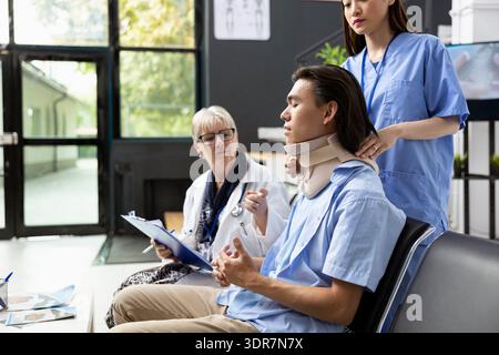 Young adult patient sits unwell in hospital waiting area wearing cervical collar. Doctor and nurse explain medical procedures, diagnosis and upcoming treatment with professional teamwork and care. Stock Photo
