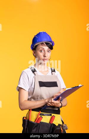 Caucasian builder holding clipboard and showing calm focus while overseeing tasks. Female contractor stands equipped with tool belt, representing project management in construction industry. Stock Photo