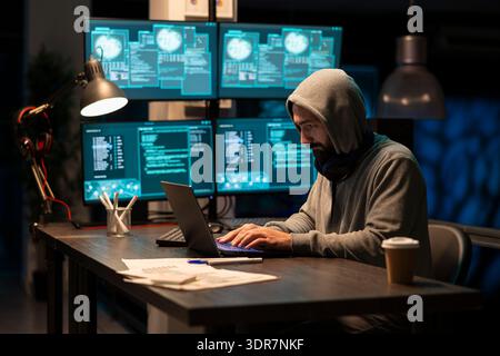 White male developer wearing hoodie and headphones concentrates on laptop screen, writing secure scripts. Freelance programmer works at night, monitoring networks and ensuring cybersecurity safety. Stock Photo