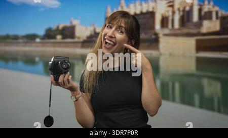 Young woman holding retro camera and pointing finger to her smile on an old town street by a waterfront building; joy travel memory. Stock Photo