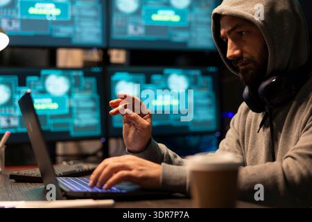 Adult male developer with focused expression debugs network systems during night shift in office. White man wearing hoodie, works on laptop, inputting code and handling cloud computing tasks. Stock Photo