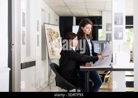 Young professionals at office desk comparing printed architectural drawings and structural plans. Women architects collaborate on project development using scale model for design accuracy and planning Stock Photo