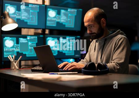 Dedicated male programmer in sweatshirt reviews system data and writes coding scripts on laptop. Focused developer improving cloud computing protection to prevent cybercrime during night shifts. Stock Photo