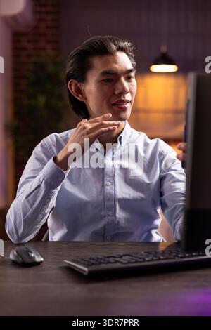 Professional man using smartphone for late night video meeting, explaining marketing strategy with hand gestures. Remote asian freelancer connecting virtually with client across time zones. Stock Photo