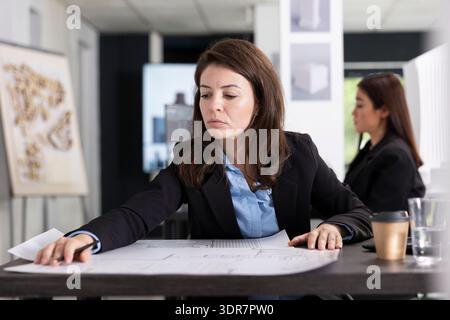 Female architect reviews blueprints on desk and prepares for presentation. Young architectural designer examines project layouts, demonstrating concentration, technical skill and attention to detail. Stock Photo