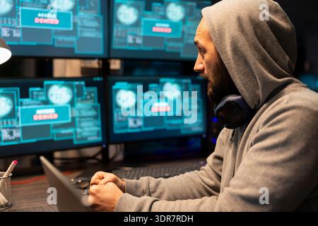 Caucasian software developer sits in dark office, monitoring encrypted data across several screens. Adult man with hoodie, concentrates on debugging network issues and enhancing information safety. Stock Photo