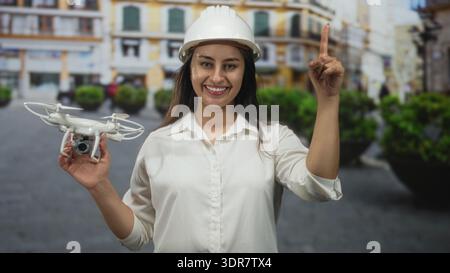 Woman engineer wearing hardhat holding white drone camera points finger up on city street; urban planning confidence. Stock Photo