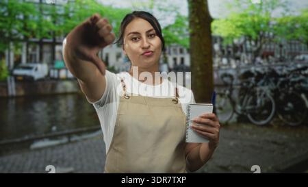 Woman waitress in apron holding notebook shows thumbs down on street in amsterdam; disapproval service feedback. Stock Photo