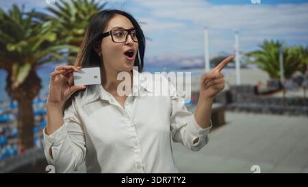 Woman holding white creditcard near face and points finger toward beach promenade with palm trees while wearing glasses and open mouth; surprise savin Stock Photo