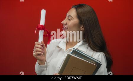 Young hispanic woman holds diploma with red ribbon and stack of books in studio; proud achievement. Stock Photo