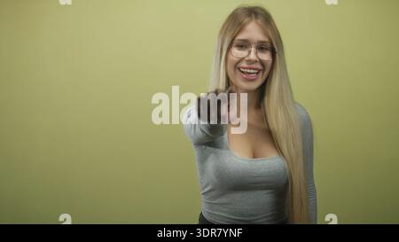 Woman points finger at camera while smiling and showing cleavage in studio, wearing glasses and grey fitted top with long blonde hair; joy playfulness Stock Photo