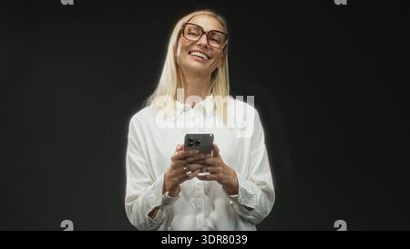 Woman middle aged blonde wearing glasses and white shirt holding smartphone with visible hands texting and smiling in studio; calm contentment. Stock Photo