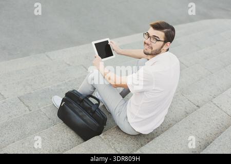 Man Uses Tablet While Sitting on Steps With Bag Beside Him Stock Photo