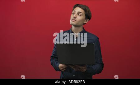 Man holding laptop with finger to chin in studio, looking up while evaluating ideas amid a red backdrop and shifting poses across frames; thoughtful f Stock Photo